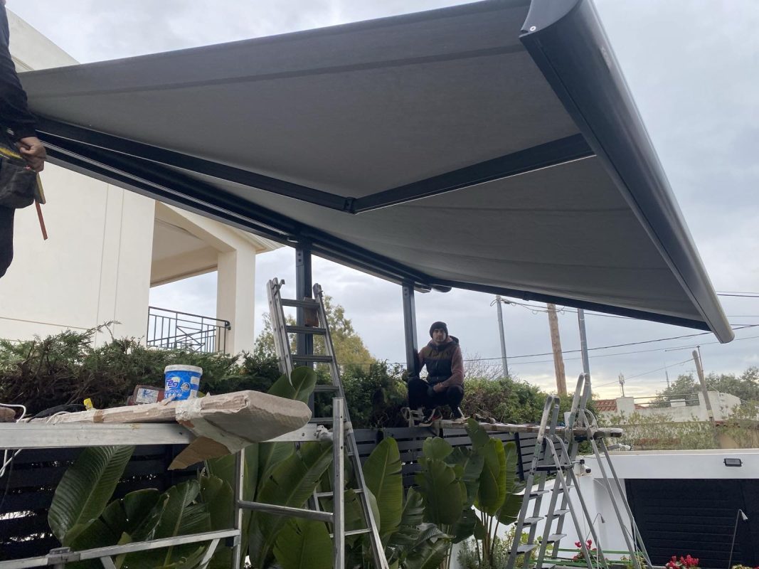 Τεχνικοί εγκαθιστούν τέντα κασετίνα με ηλεκτρικό μοτέρ σε εξωτερικό χώρο κατοικίας στη Βάρη, χρησιμοποιώντας σκάλες και εξειδικευμένα εργαλεία.