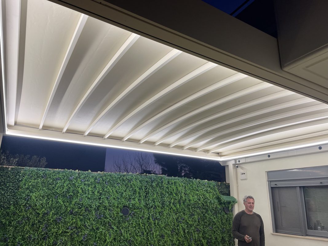 Flat τεντοπέργκολα με LED φωτισμό dimmable, φωτισμένη το βράδυ σε έναν καλαίσθητο εξωτερικό χώρο με φυτά στον τοίχο.