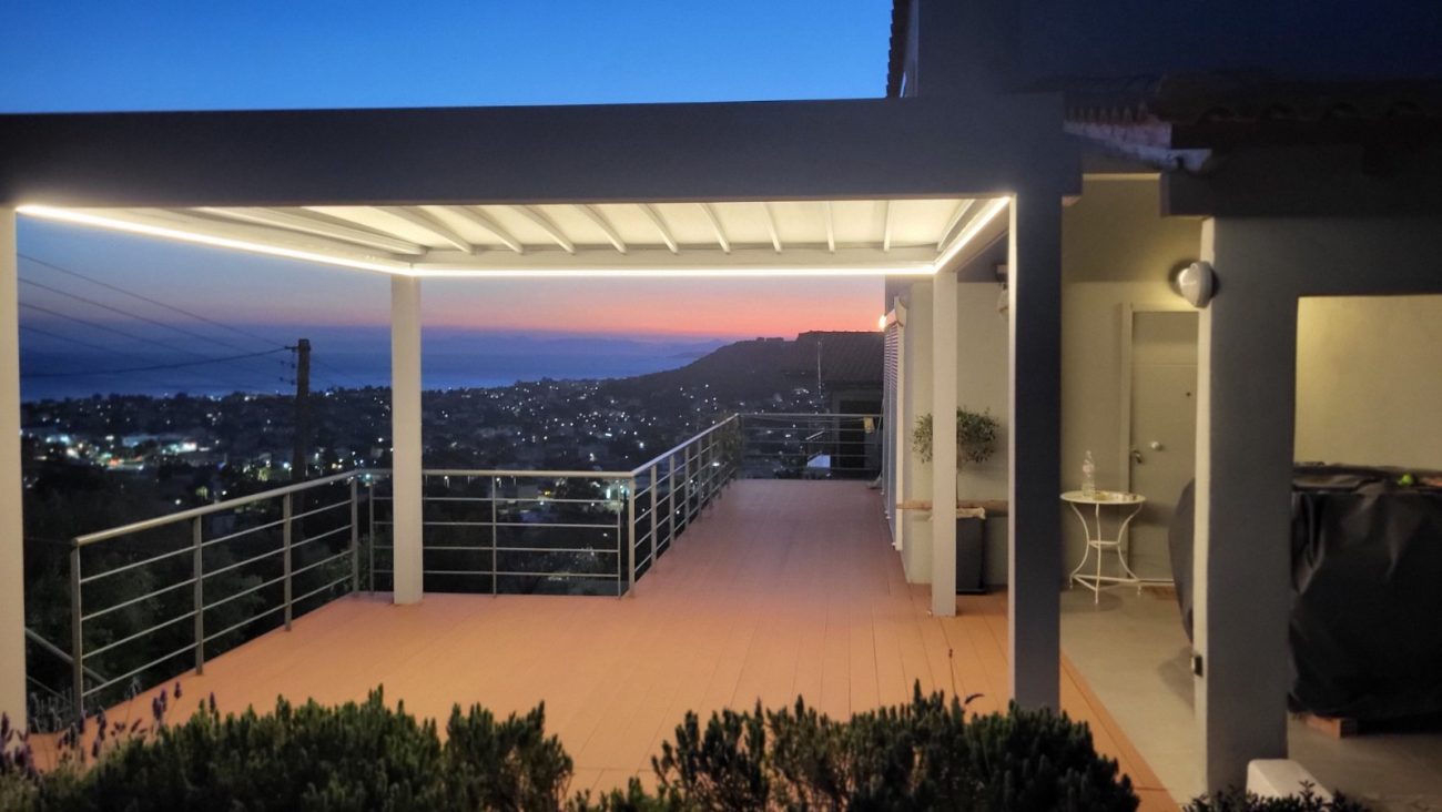 Flat τεντοπέργκολα με περιμετρικό LED φωτισμό, φωτισμένη το βράδυ σε βεράντα με μεταλλικά κάγκελα και θέα στη θάλασσα.