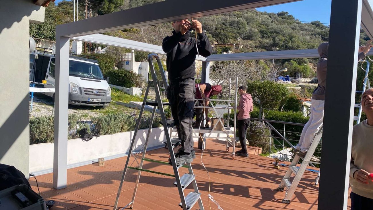 Επαγγελματίες τεχνικοί συναρμολογούν flat τεντοπέργκολα, τοποθετώντας μεταλλικά δοκάρια σε εξωτερικό χώρο.